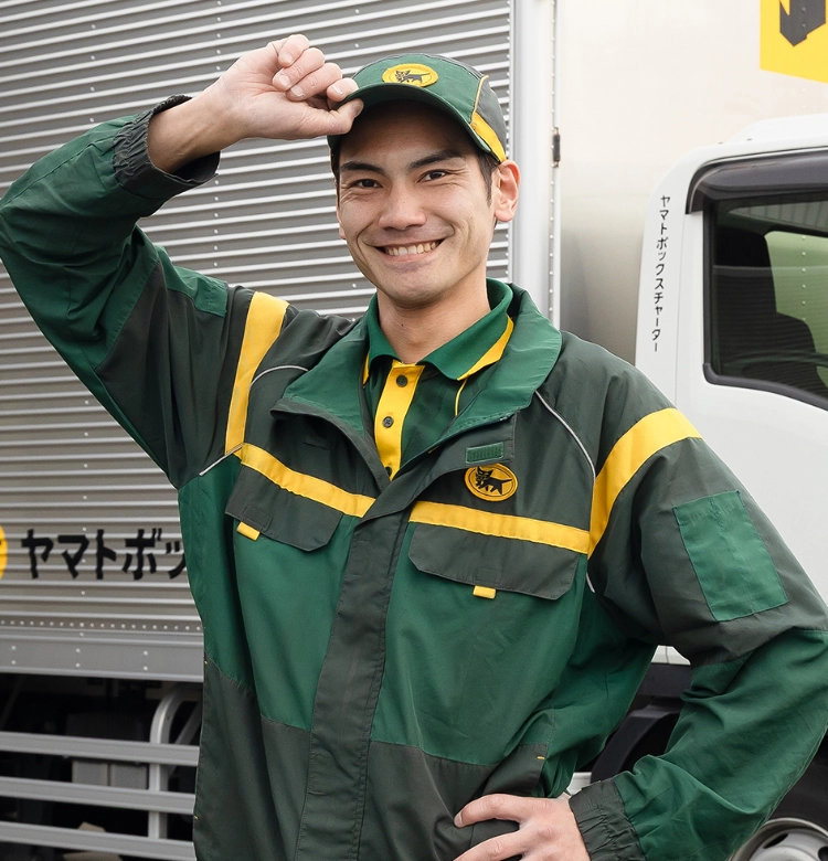ヤマトボックスチャーターのトラックの前で、緑と黄色のユニフォームを着てキャップに手を添え、満面の笑みを浮かべる男性ドライバー
