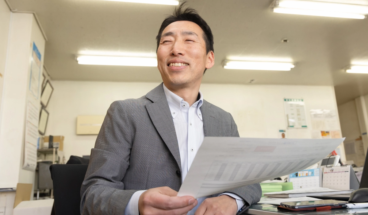 I.A.さんインタビューキービジュアル。デスクで資料を手に持ち、柔らかな笑顔で横を向く法人営業担当のI.A.さん