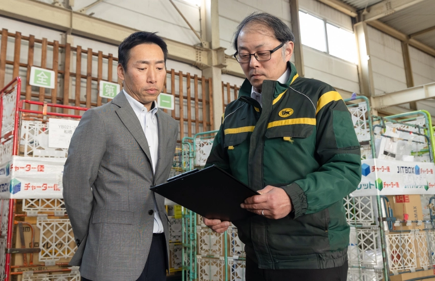 物流倉庫にて、作業服を着た現場スタッフとバインダーの資料を確認しながら真剣に打ち合わせをする法人営業のI.A.さん
