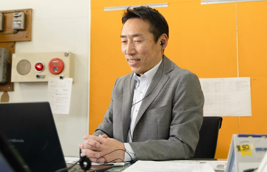 イヤホンを装着し、デスクのノートPCに向かって笑顔でリモート打ち合わせを行う法人営業のI.A.さん