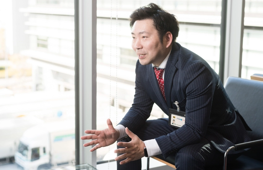 窓の外にトラックが並ぶ開放的なオフィスロビーで、身振り手振りを交えて熱心に話すS.T.さん