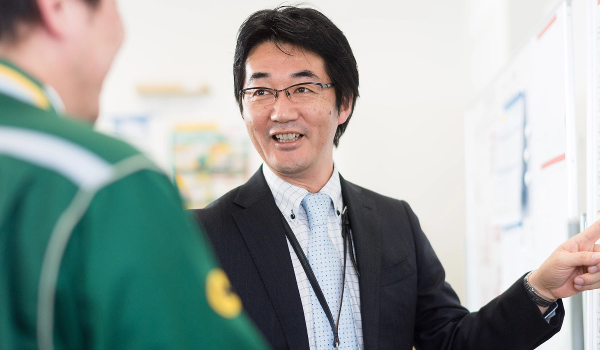 N.K.さんインタビューキービジュアル。現場スタッフとコミュニケーションをとるN.K.さん。ホワイトボードの前で業務を確認する様子
