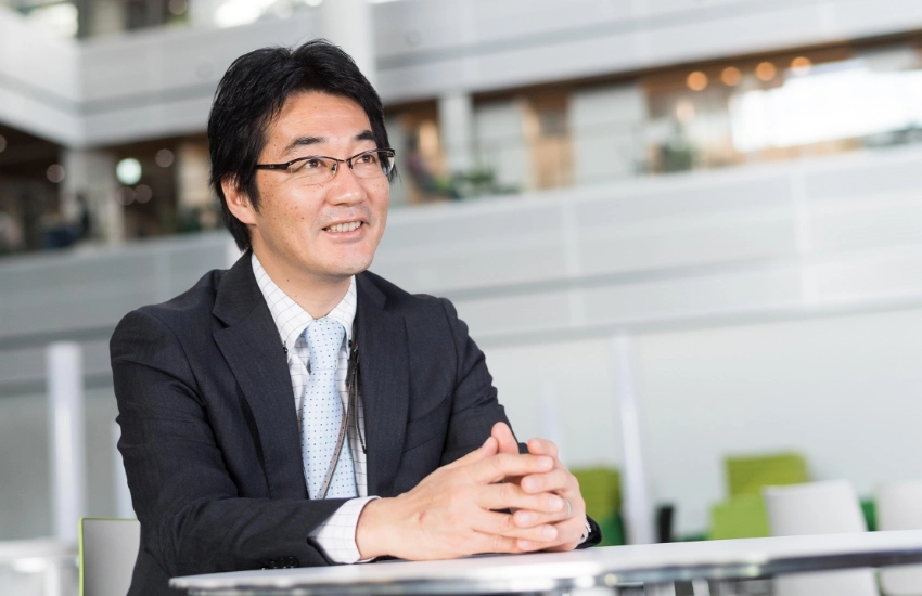 広々とした明るいオフィスロビーのデスクで、手を組みながら穏やかな表情で話をするN.K.さん