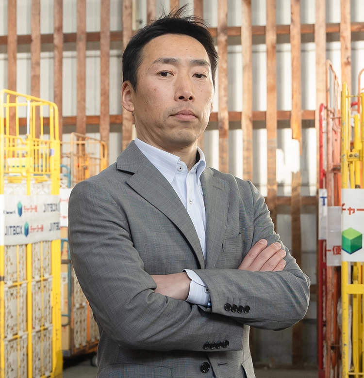 広い倉庫内でチャーター便のカゴ台車を背景に腕組みをして凛々しい表情で立っている法人営業の男性社員